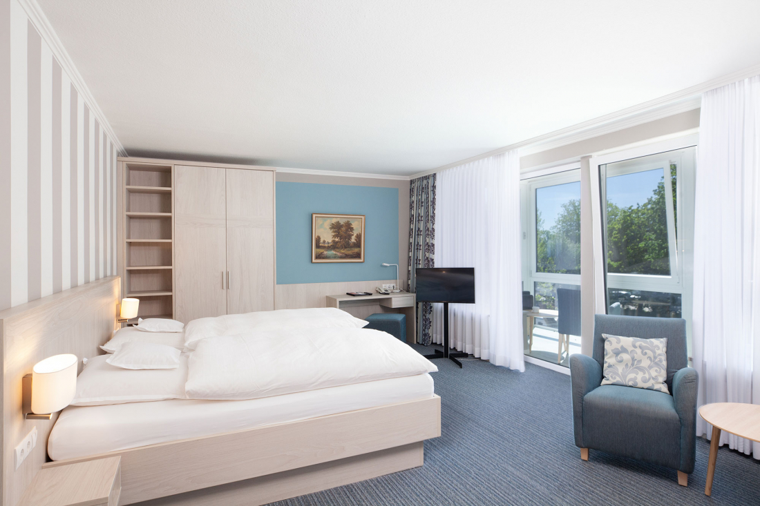 Ein helles Hotelzimmer mit einem Doppelbett, Sessel, Schreibtisch, Fernseher, großem Kleiderschrank und einem Fenster mit durchsichtigen Vorhängen mit Blick ins Grüne. Die Einrichtung ist hell mit blauen und weißen Tönen und einem Gemälde an der Wand.