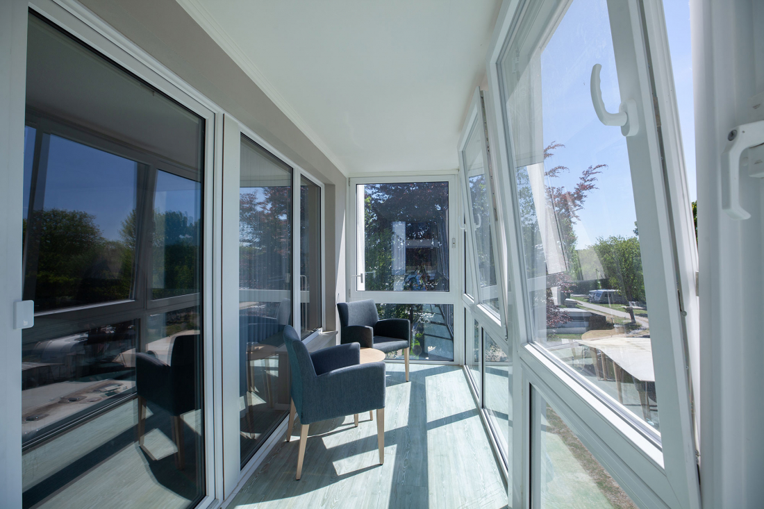 Ein heller, geschlossener Balkon mit großen Glasfenstern, zwei modernen grauen Stühlen, einem kleinen runden Tisch und Sonnenlicht, das auf den hellen Holzboden fällt. Draußen sind Bäume und Grünflächen zu sehen.