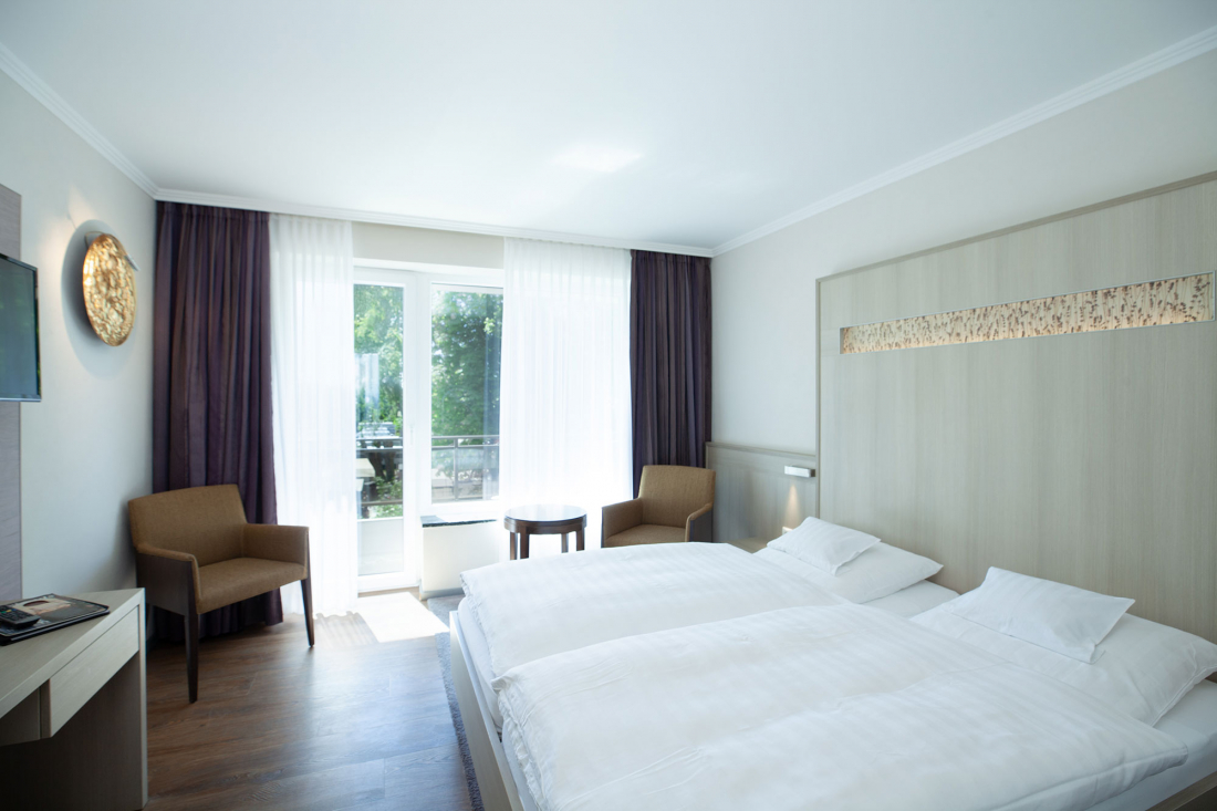 Helles Hotelzimmer mit einem großen Bett, zwei braunen Stühlen, einem kleinen runden Tisch, einem Schreibtisch mit Fernseher und einem Fenster mit durchsichtigen Vorhängen, die natürliches Licht hereinlassen. Die Einrichtung ist modern und minimalistisch.