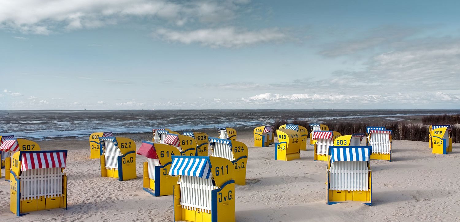 Gelbe Strandkörbe mit gestreiften blauen oder roten Schirmen sind an einem Sandstrand mit Blick auf das Meer unter einem teilweise bewölkten Himmel angeordnet. Jeder Stuhl trägt eine Nummer und die Initialen J.v.G.