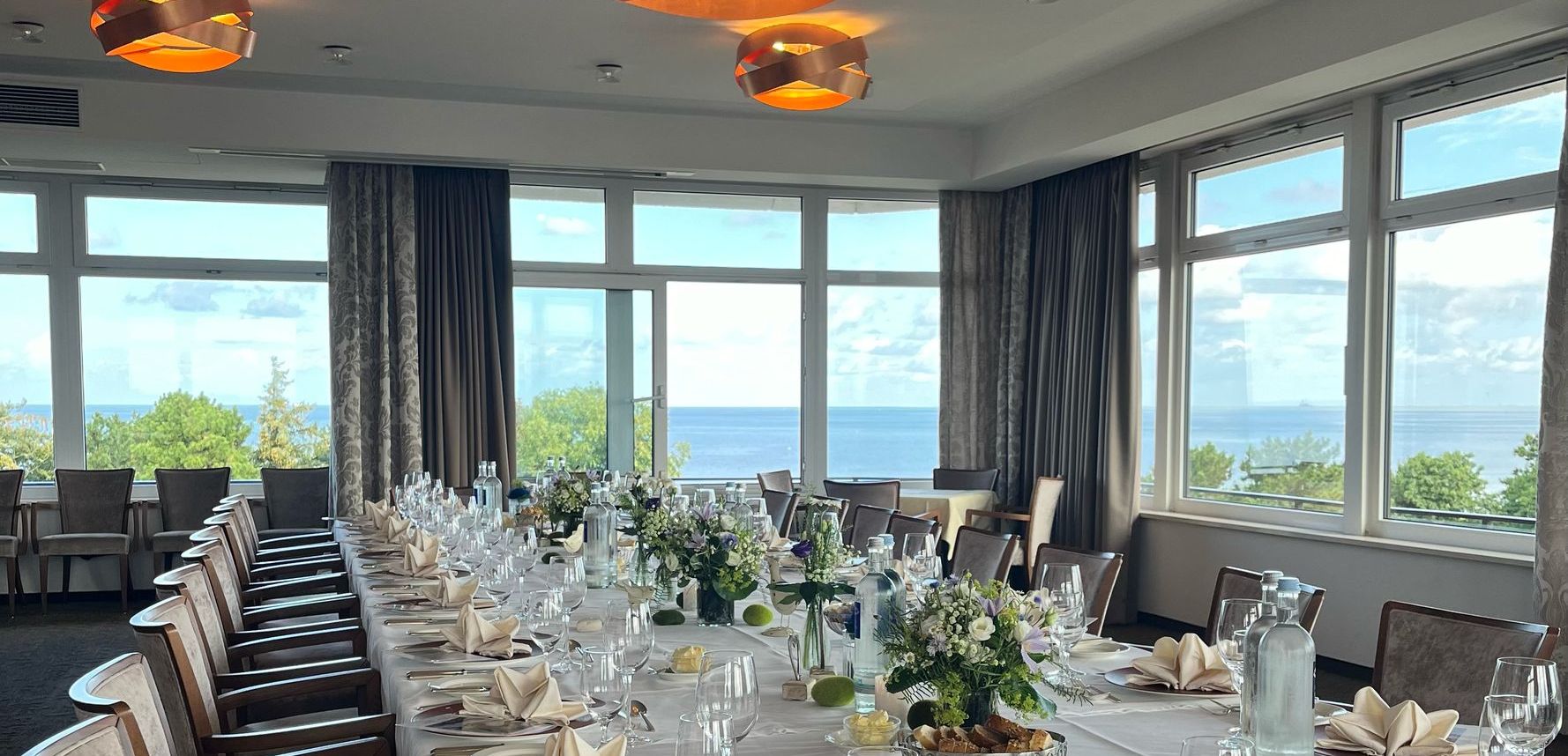Ein langer, für eine elegante Veranstaltung gedeckter Tisch mit weißen Tischtüchern, gefalteten Servietten, Gläsern, Blumenschmuck und großen Fenstern, die einen malerischen Blick auf Bäume und die Nordsee unter einem wolkenfreien Himmel bieten.