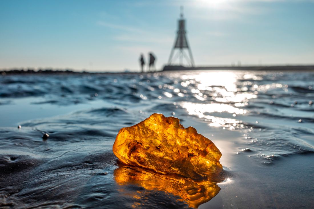 Ein großes Stück Bernstein liegt im nassen Watt an der Küste und glitzert im Sonnenlicht. Im Hintergrund gehen zwei unscharfe Gestalten unter strahlend blauem Himmel auf die Kugelbake zu.