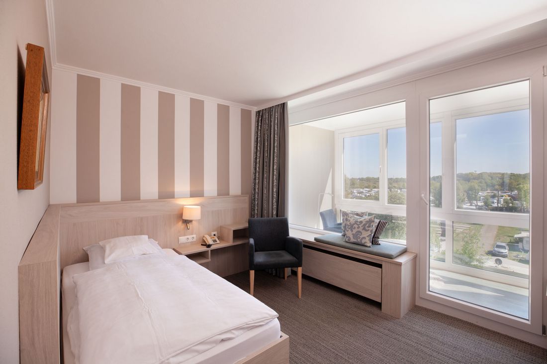 Ein helles, modernes Hotelzimmer mit einem Einzelbett, einer gestreiften Akzentwand, einem Nachttisch, einem Sessel und einem großen Fensterplatz mit Blick auf einen malerischen Außenbereich. Das Zimmer ist von natürlichem Licht durchflutet.