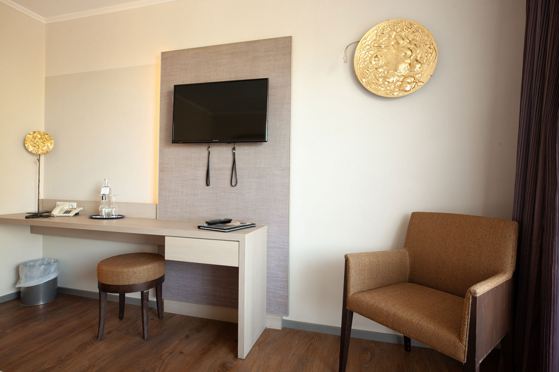 Ein moderner kleiner Ablagebereich im Doppelzimmer mit einem an der Wand montierten Fernseher, einem Schreibtisch mit Hocker, Telefon, Wasserflasche und Notizblock, einem braunen Sessel und zwei goldenen dekorativen Wandtafeln.