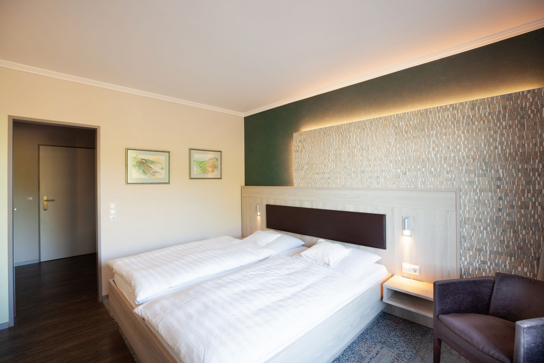 Ein modernes Hotelzimmer mit einem Doppelbett, weißer Bettwäsche, einer gemusterten Akzentwand, zwei Gemälden, einem Nachttisch und einem violetten Sessel neben dem Bett. Das Zimmer ist warm beleuchtet und hat eine Tür auf der linken Seite.