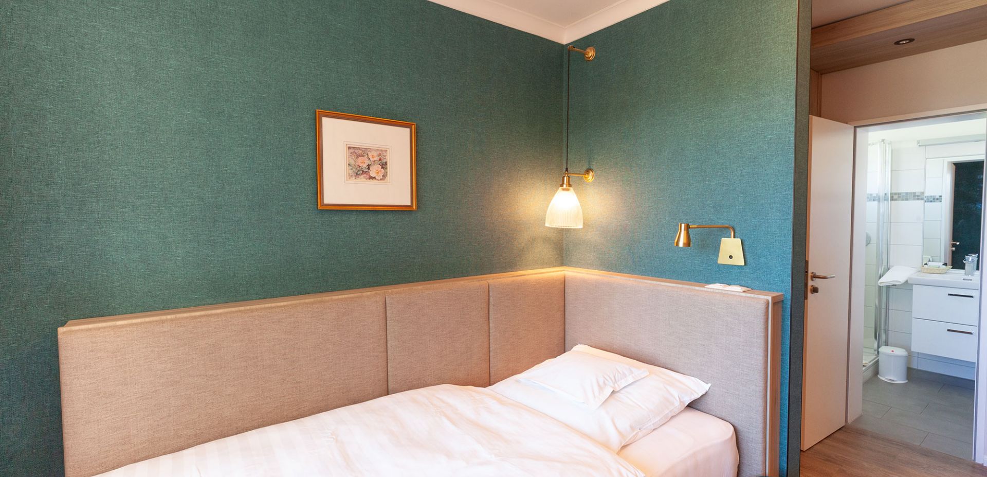 Ein kleines, modernes Hotelzimmer mit tealfarbenen Wänden, einem Einzelbett mit weißer Bettwäsche, einer Wandlampe, gerahmten Kunstwerken und einer offenen Tür, die in ein helles Badezimmer führt.