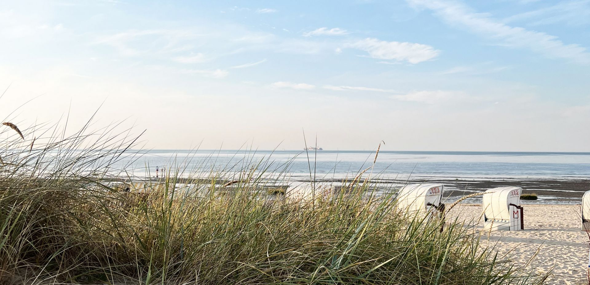 Im Vordergrund hohes Strandgras, dazu weiße Strandkörbe am Strand in Duhnen. Im Hintergrund befindet sich die Nordsee bei ablaufend Wasser und blauem Himmel; am fernen Horizont ist ein Schiff zu sehen.