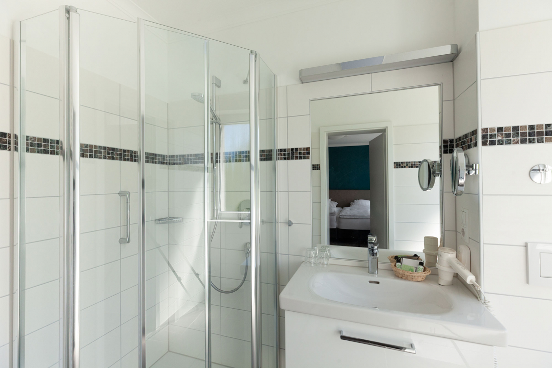 Modernes Badezimmer mit weiß gefliesten Wänden, einer verglasten Eckdusche, einem weißen Waschbecken mit einem Spiegel darüber und einem Haartrockner auf dem Tresen. Eine offene Tür gibt den Blick auf ein Schlafzimmer im Hintergrund frei.