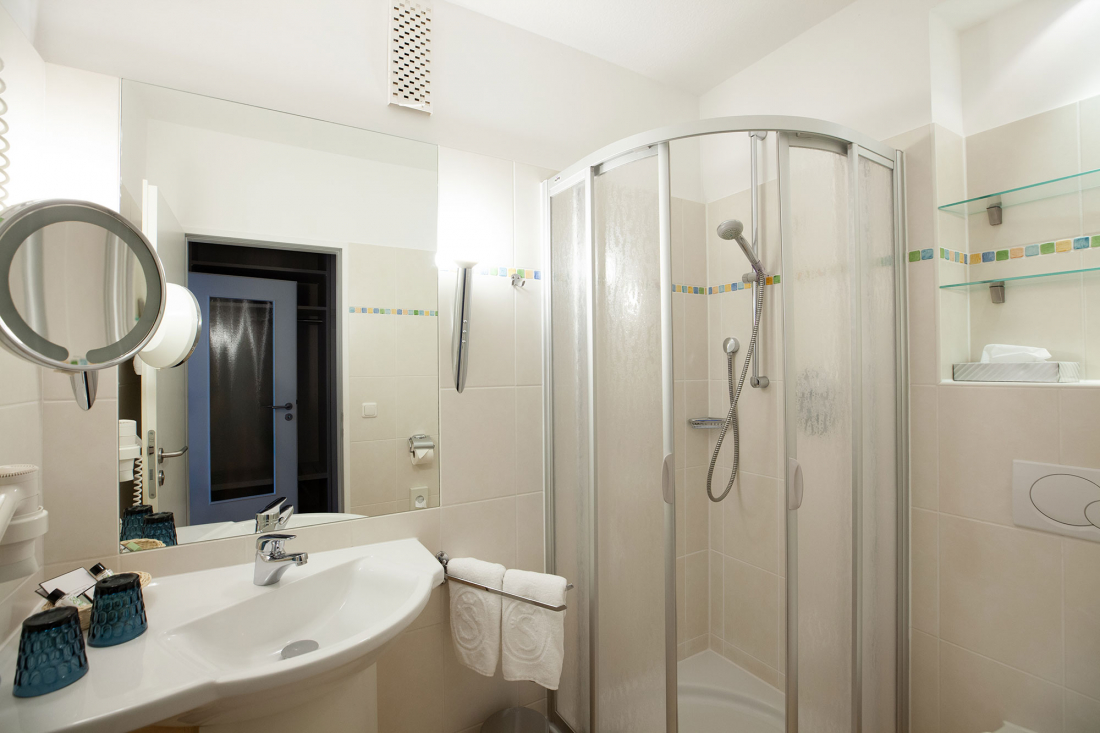 Ein modernes, sauberes Badezimmer mit einer Glasduschkabine, einem Waschbecken mit Spiegel, Wandregalen, Handtüchern und blauen Toilettenbechern auf dem Tresen. Die Wände sind in hellem Beige gefliest.