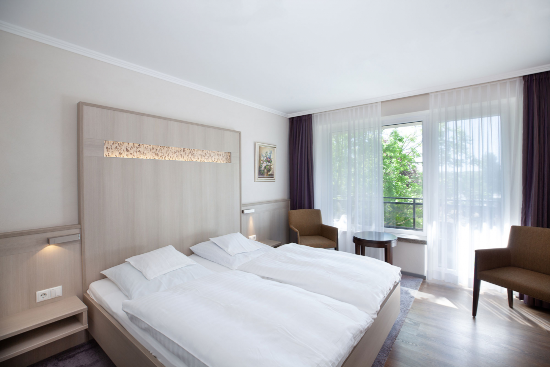 Ein helles, modernes Hotelzimmer mit zwei zusammengeschobenen Einzelbetten, weißem Bettzeug, Holzkopfteil, zwei braunen Stühlen an einem runden Tisch, einem großen Fenster mit durchsichtigen Vorhängen und einem Gemälde an der Wand.