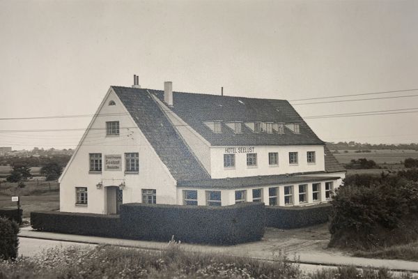 Historisches Schwarz-Weiß Foto vom ursprünglichen großen, zweistöckigen Gebäudes mit steilem Dach, das die Bezeichnung Hotel Seelust trägt. Das Hotel ist von Hecken und Feldern umgeben, und vor dem Gebäude verläuft eine Straße.