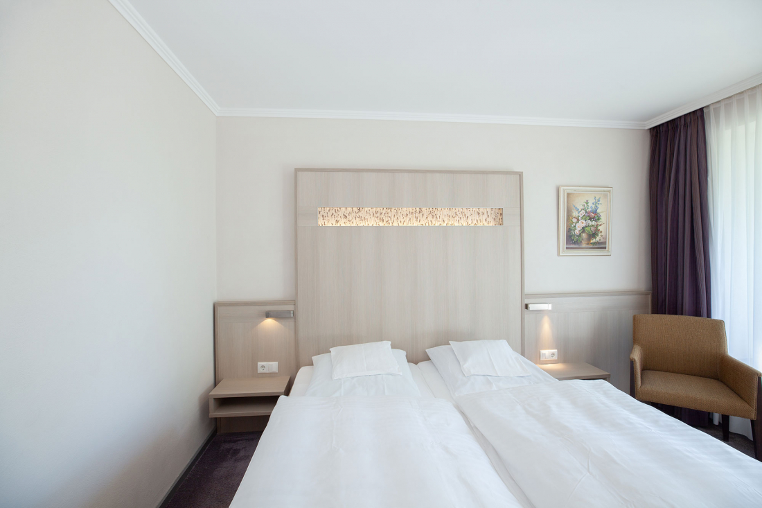 Ein Doppelzimmer mit Doppelbett, weißem Bettzeug, einem hellen Holzkopfteil mit eingebauten Lampen, einem Nachttisch auf jeder Seite, einem Sessel, einem gerahmten Bild und einem großen Fenster mit durchsichtigen Vorhängen.