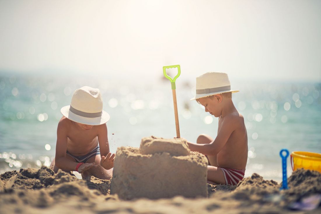 Zwei kleine Jungen mit Strohhüten bauen an einem sonnigen Strand eine Sandburg. Ein Junge klopft Sand, während der andere sich auf die Burg konzentriert, aus der eine grüne Schaufel herausragt. Im Hintergrund ist das Meer zu sehen.