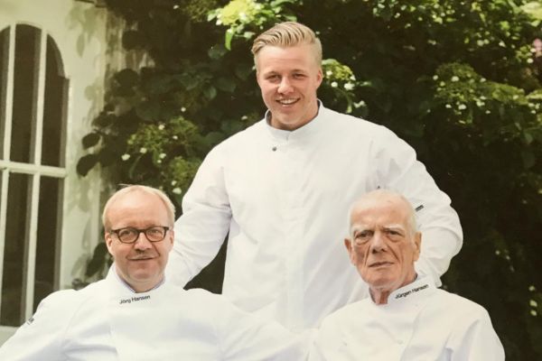 Nils Hansen (stehend) hinter den auf einer braunen Holzbank sitzenden Jörg Hansen und Jürgen Hansen im Garten.