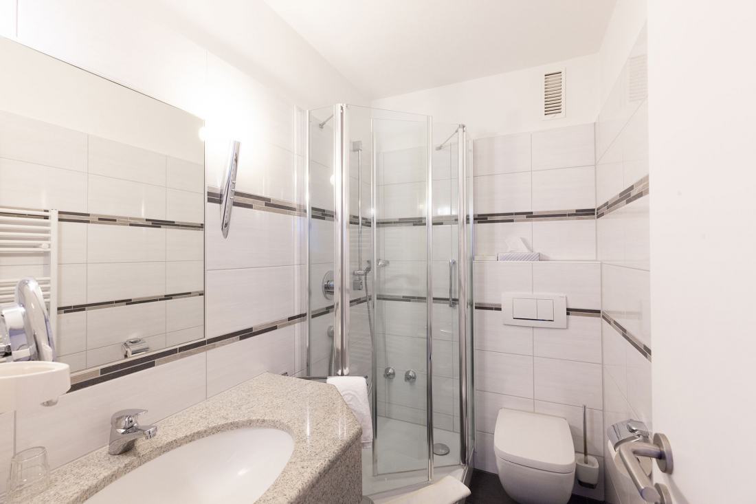 Modernes weißes Badezimmer mit gefliesten Wänden, einer Eckdusche mit Glasabtrennung, einer an der Wand befestigten Toilette, einem Waschbecken mit Granitarbeitsplatte und einem großen Spiegel über dem Waschbecken.