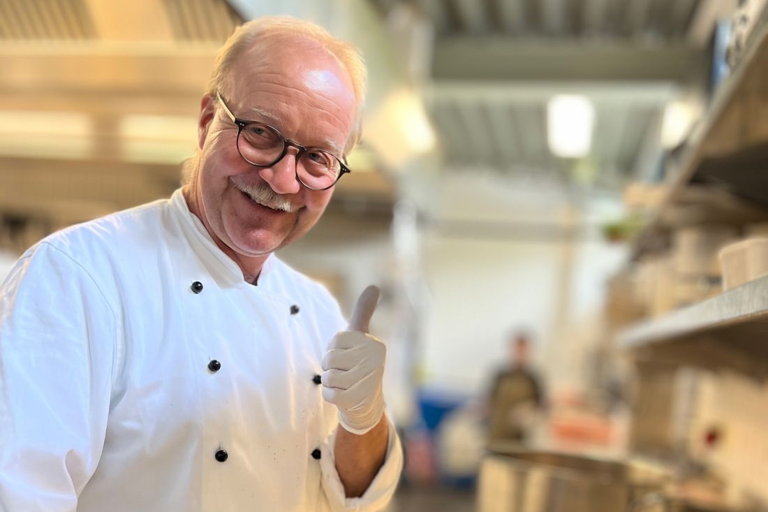 Jörg hansen mit Brille und Schnurrbart zeigt in einer belebten Küche den Daumen nach oben. Er trägt eine weiße Kochjacke und Einweghandschuhe. Der Hintergrund ist unscharf und zeigt Küchengeräte und eine weitere Person.