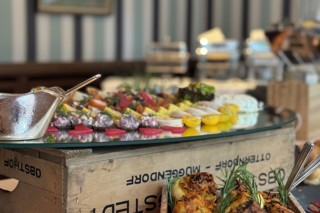 Nahaufnahme eines Buffettisches mit einer Auswahl an bunten Vorspeisen und Käse auf einer Glasfläche, die auf einer Holzkiste ausgestellt ist, mit Servierutensilien und einem verschwommenen Hintergrund.