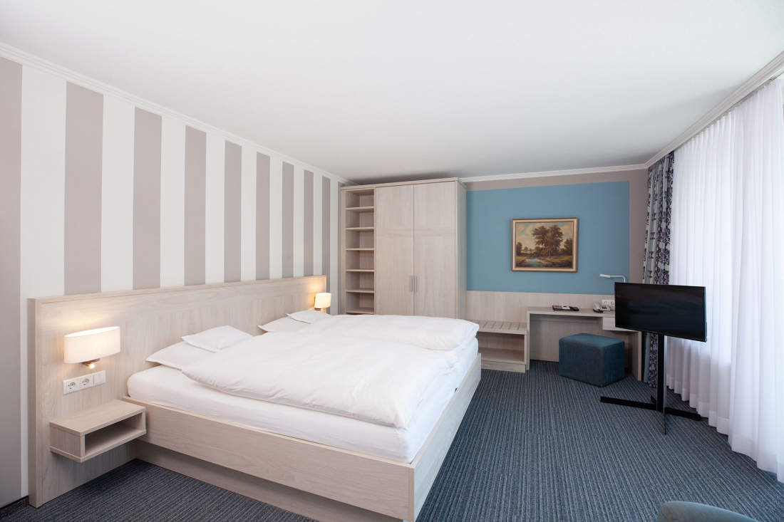 Ein modernes Hotelzimmer mit einem Doppelbett, gestreifter Tapete, Nachttischlampen, einem Kleiderschrank, einem Schreibtisch mit Fernseher und einem großen Fenster mit durchsichtigen Vorhängen, durch das natürliches Licht fällt.