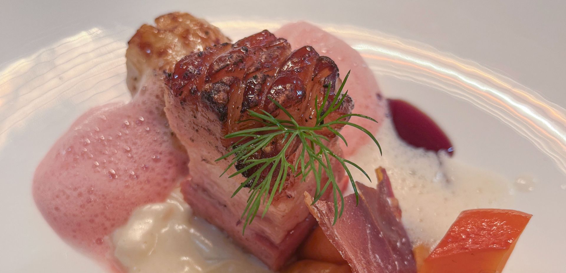 Nahaufnahme eines Gourmetgerichts mit einem gebratenen Stück Fleisch, das mit frischem Dill, rosa Schaum, cremiger weißer Soße, einer Scheibe gepökeltem Fleisch und Stücken von rotem Gemüse kunstvoll auf einem weißen Teller angerichtet ist.