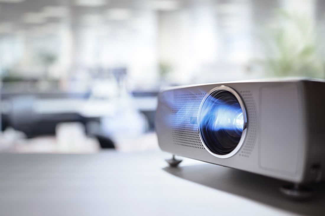 Nahaufnahme eines Digitalprojektors auf einem Tisch in einem hellen, modernen Büro, dessen Linse blaues Licht ausstrahlt und dessen Hintergrund aus Schreibtischen und Lichtern verschwommen ist.