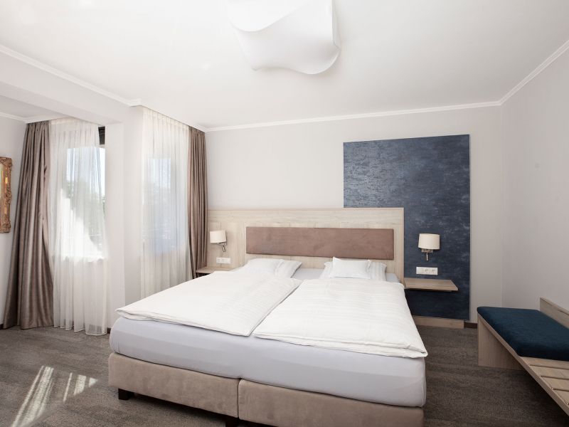 Ein modernes Hotelzimmer des Drei-Raum-Apartments mit einem großen Doppelbett, weißem Bettzeug, zwei Nachttischen mit Lampen, einer blauen Akzentwand, großen Fenstern mit Vorhängen und einer Bank an einer Wand.
