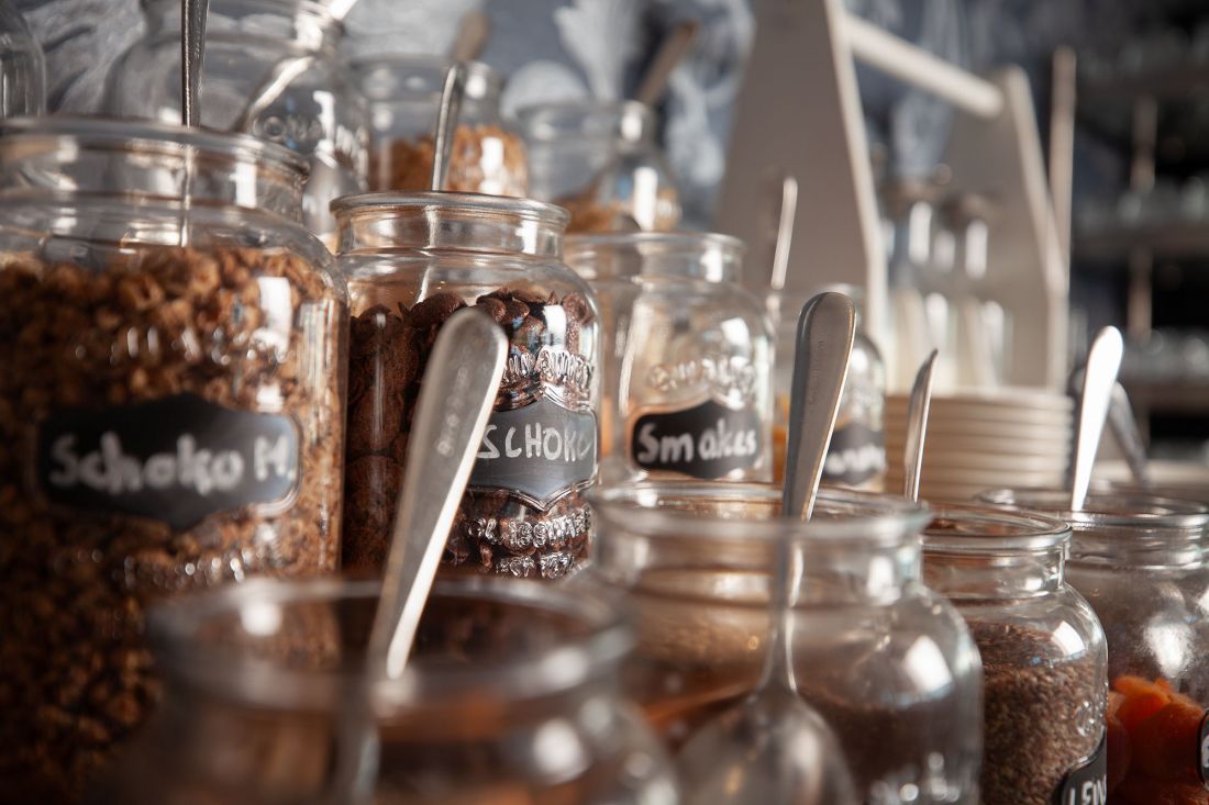 Eine Reihe von Gläsern mit verschiedenen Cerealien und Toppings, jedes mit einem Löffel und einem kleinen Kreidetafel-Etikett, die auf einer Frühstückstheke oder einem Buffet stehen.