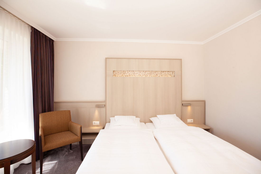 Ein helles, modernes Hotelzimmer mit Doppelbett, weißem Bettzeug, einem hölzernen Kopfteil, einem braunen Sessel, einem kleinen runden Tisch und einem großen Fenster mit durchsichtigen und dunklen Vorhängen.