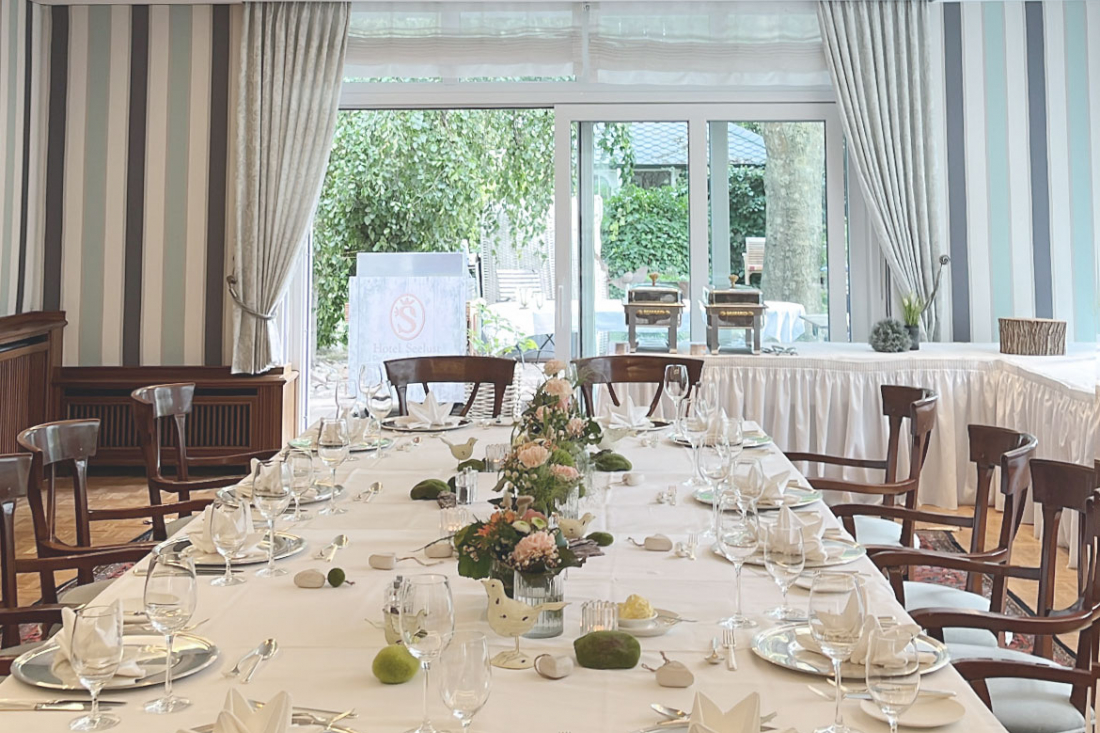 Tagungsraum 1 "Gartenbrise" mit gestreifter Tapete, großen Fenstern, Holzstühlen und einem langen, mit weißer Bettwäsche, Gläsern und Servietten gedeckten Tisch, bereit für ein formelles Essen. Durch die Fenster ist das Grün draußen zu sehen.