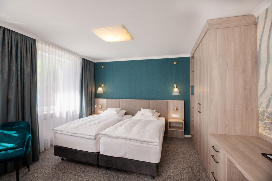 Ein modernes Hotelzimmer mit zwei aneinander geschobenen Einzelbetten, tealfarbener Akzentwand, hellen Holzmöbeln, tealfarbenem Stuhl, großem Kleiderschrank und einem Fenster mit Verdunkelungsvorhängen, die natürliches Licht hereinlassen.