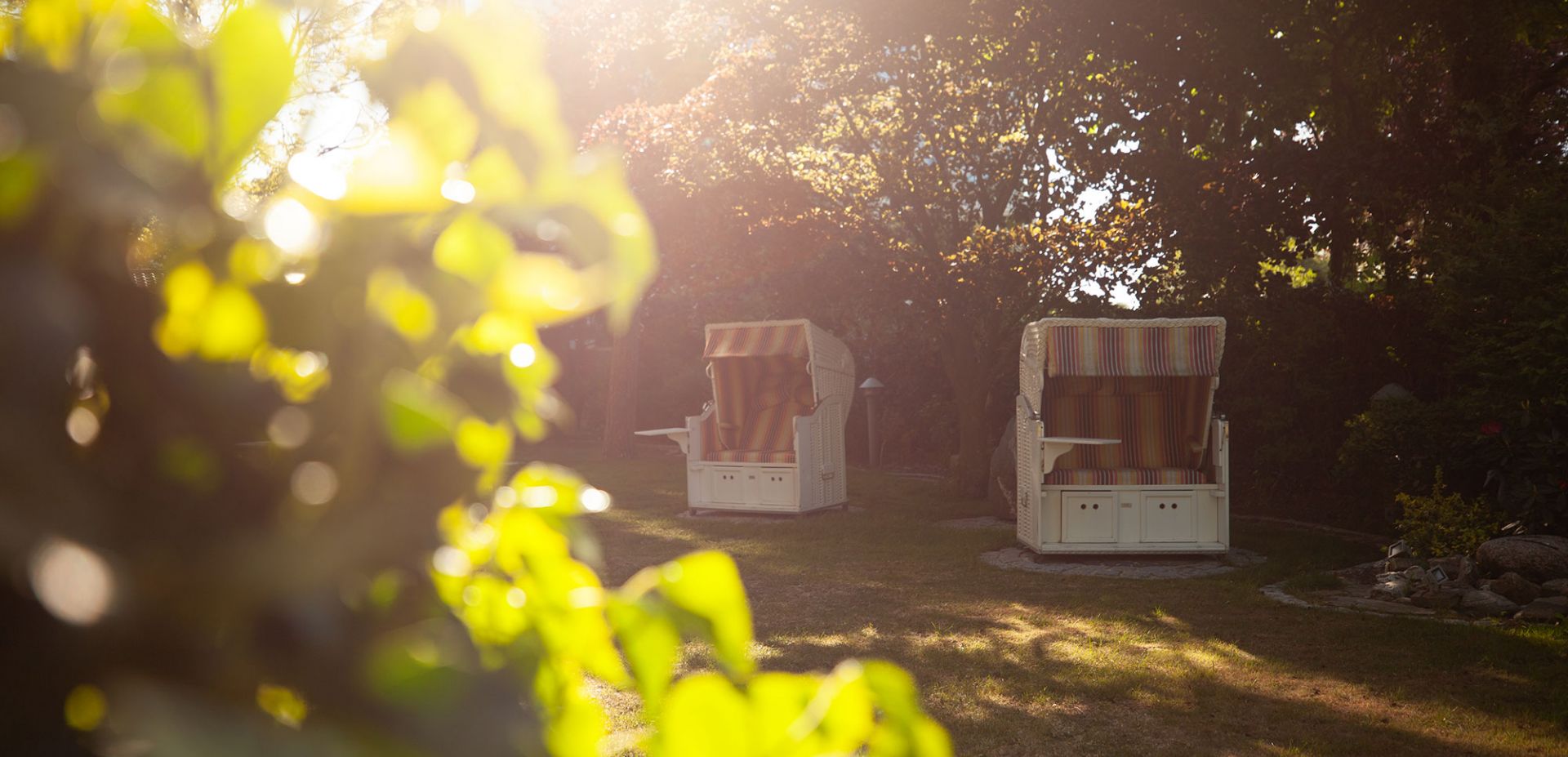 Das Sonnenlicht dringt durch die belaubten Äste und beleuchtet zwei gestreifte Strandkörbe mit Baldachin, die auf dem Gras in einem schattigen, ruhigen Garten stehen. Die Atmosphäre ist heiter und einladend.