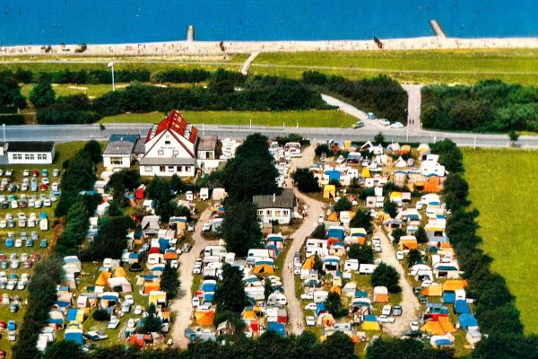 Luftaufnahme des vollen Campingplatzes Seelust mit farbenfrohen Zelten und Wohnwagen, die entlang kurvenreicher Straßen aufgestellt sind, begrenzt durch grüne Felder, des Hotel Seelust, einen Parkplatz und der Nordsee im Hintergrund.