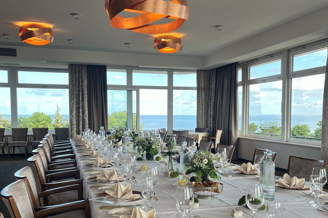 Ein langer, für eine elegante Veranstaltung gedeckter Tisch mit weißen Tischtüchern, gefalteten Servietten, Gläsern, Blumenschmuck und großen Fenstern, die einen malerischen Blick auf Bäume und die Nordsee unter einem wolkenfreien Himmel bieten.