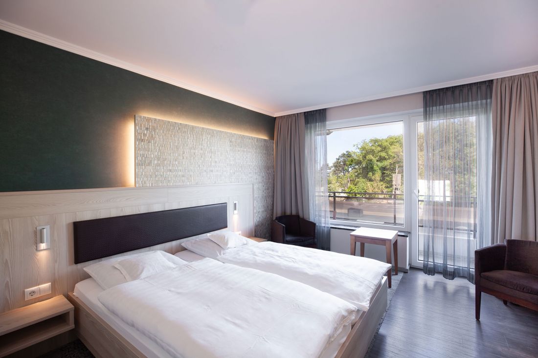 Ein modernes Doppelzimmer mit Doppelbett, weißem Bettzeug, einem hölzernen Kopfteil mit eingebauten Lampen, einer dunkelgrünen Akzentwand, einem großen Fenster mit Verdunkelungsvorhängen, Stühlen und einem kleinen Tisch.