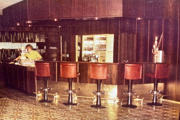 Ein Barkeeper arbeitet hinter einer Bar aus dunklem Holz mit vier rot gepolsterten Barhockern davor. Der Barbereich ist mit Flaschen und Gläsern bestückt, der gemusterte Fliesenboden und die gedämpfte Beleuchtung sorgen für eine Vintage-Atmosphäre.