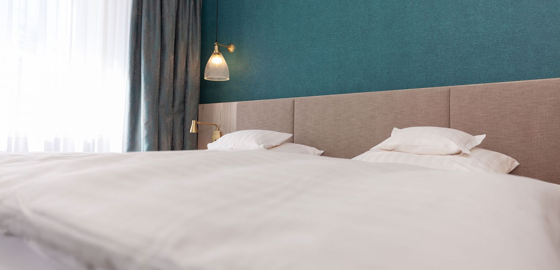 Ein ordentlich gemachtes Bett mit weißer Bettwäsche und Kissen in einem modernen Zimmer mit einer tealfarbenen Akzentwand, einem beigen Kopfteil und einer goldenen Wandlampe neben einem Fenster mit weißen Vorhängen und dunklen Gardinen.