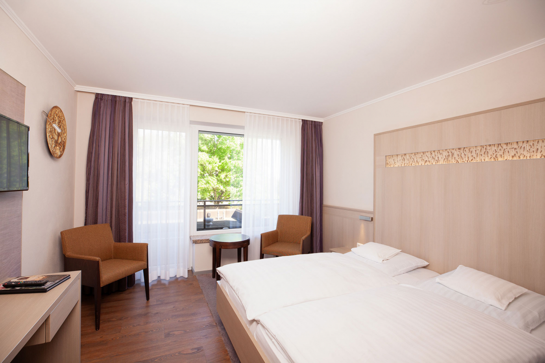 Ein helles Hotelzimmer mit einem Doppelbett, zwei braunen Stühlen, einem kleinen runden Tisch an einem Fenster mit durchsichtigen Vorhängen, einem Fernseher an der Wand und hellen Holzmöbeln. Draußen sind grüne Bäume zu sehen.