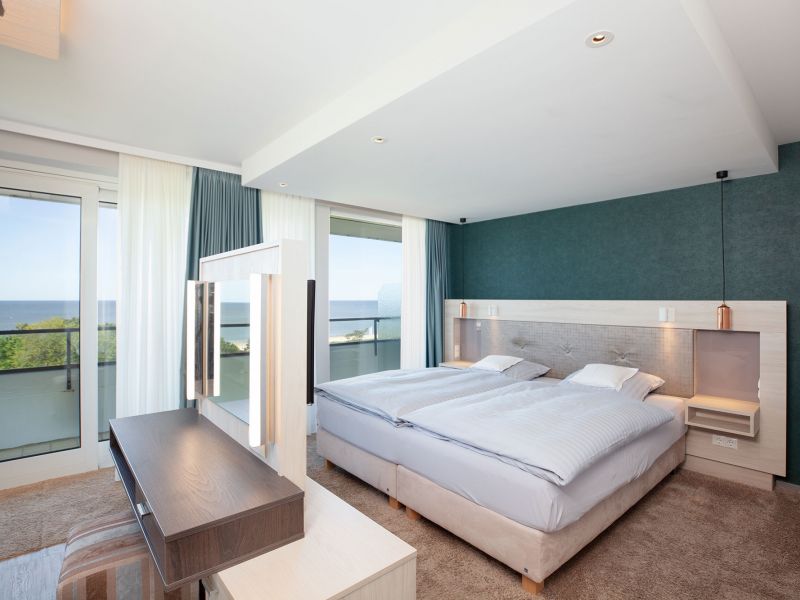 Modernes Zwei-Raum-Suite mit zwei Einzelbetten, einem großen Fenster mit durchsichtigen Vorhängen, einem Balkon, einem Schreibtisch mit Spiegel und Blick auf die Nordsee..