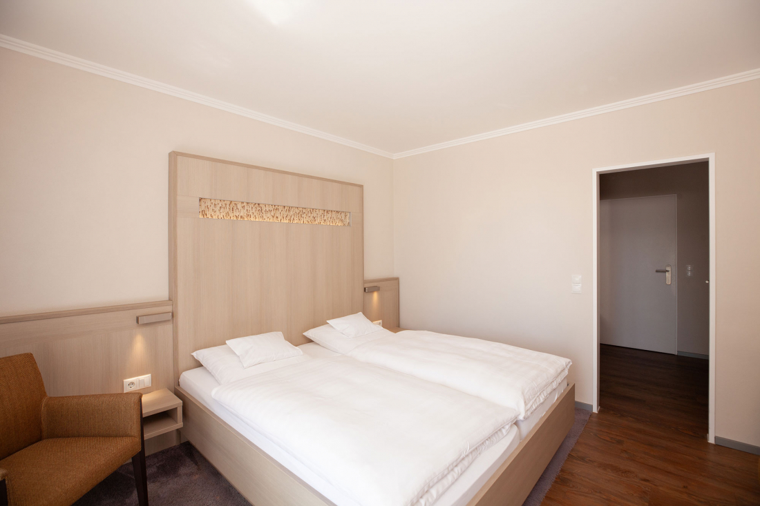Ein minimalistisches Doppelzimmer mit zwei einzelnen Matrazen im Doppelbett, weißem Bettzeug, einem beigen Kopfteil, einem braunen Sessel, Holzboden und neutralen Wänden. Eine offene Tür führt in den kleinen Eingangsflur.
