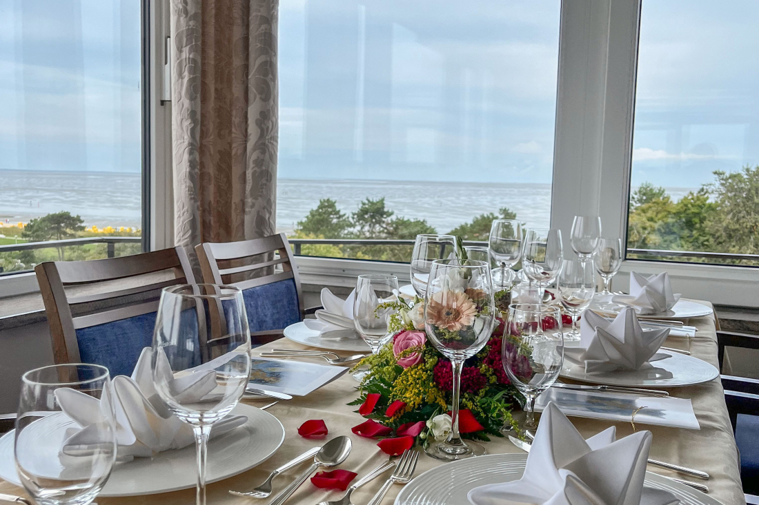 "Café zur Schönen Aussicht" Ein für eine formelle Mahlzeit gedeckter Esstisch mit gefalteten Servietten, Gläsern und Blumenschmuck, der an einem großen Fenster mit Blick auf Bäume und das Meer steht. Rote Rosenblätter sind auf dem Tisch verstreut.