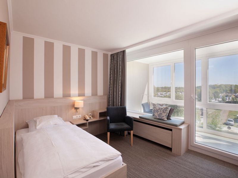 Ein helles, modernes Hotelzimmer mit einem Einzelbett, einer gestreiften Akzentwand, einem Nachttisch, einem Sessel und einem großen Fensterplatz mit Blick auf einen malerischen Außenbereich. Das Zimmer ist von natürlichem Licht durchflutet.