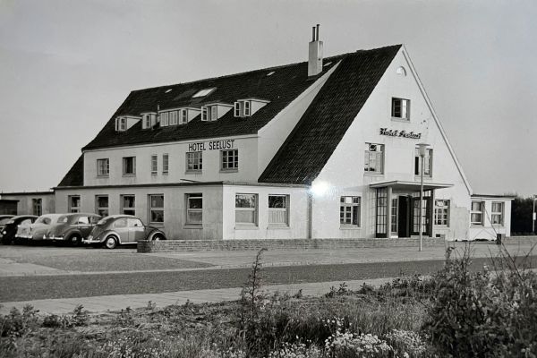 Schwarz-Weiß-Foto des zweistöckigen Hotel Seelust von 1934 mit der Aufschrift "Hotel Seelust", vor dem mehrere Oldtimer geparkt sind und das im Vordergrund von Gras umgeben ist. Die Architektur ist einfach mit einem Schrägdach und Dachgauben.