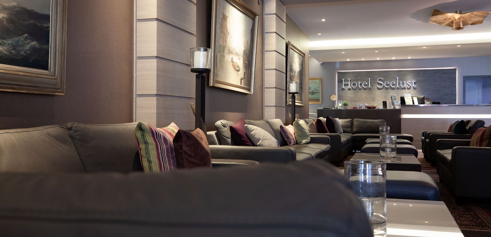 Hotellobby des Hotel Seelust mit grauen Sofas, bunten Kissen und Tischen mit Glasplatte. An den Wänden hängen Gemälde, und im Hintergrund ist die Rezeption mit einem Hotel Seelust-Schild zu sehen.