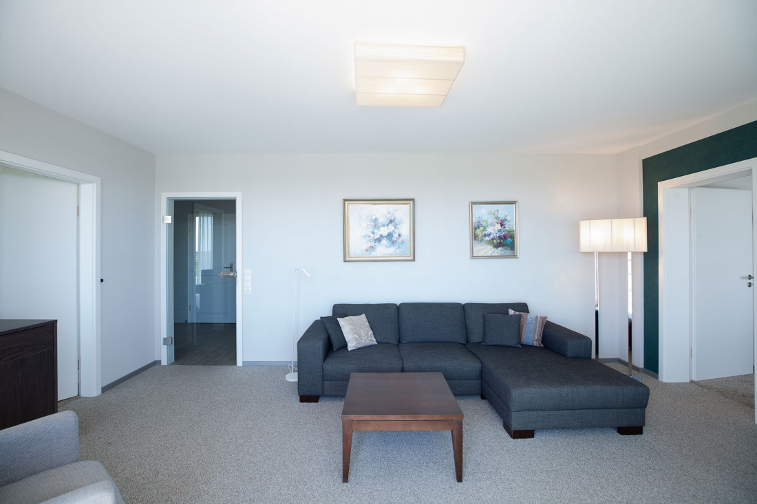 Ein modernes Wohnzimmer mit einem dunkelgrauen Sektionssofa, einem hölzernen Couchtisch, zwei gerahmten Kunstwerken an der Wand, einer Stehlampe, neutralen Wänden und Teppichboden sowie Türen, die zu anderen Räumen führen.