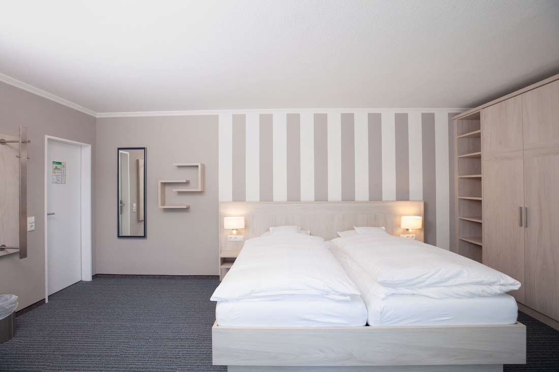 Ein modernes Hotelzimmer mit einem Doppelbett, weißer Bettwäsche, zwei Nachttischlampen, offenen Regalen, einem an der Wand befestigten Spiegel und einer gestreiften Akzentwand hinter dem Bett. Das Design ist minimalistisch mit neutralen Farben.
