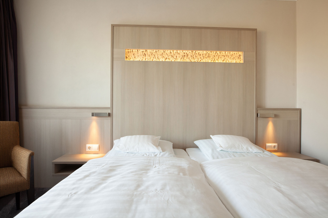 Ein Doppelzimmer mit zwei Einzelmatrazen im Doppelbett, jeweils mit weißer Bettwäsche und Kissen. Das Kopfteil hat eingebaute Lampen und eine Holzvertäfelung. Auf der linken Seite ist ein brauner Sessel teilweise sichtbar.