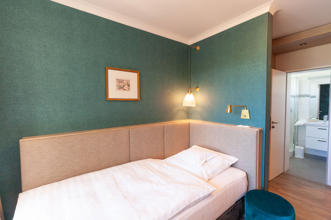 Ein kleines, modernes Hotelzimmer mit tealfarbenen Wänden, einem Einzelbett mit weißer Bettwäsche, einer Wandlampe, gerahmten Kunstwerken und einer offenen Tür, die in ein helles Badezimmer führt.