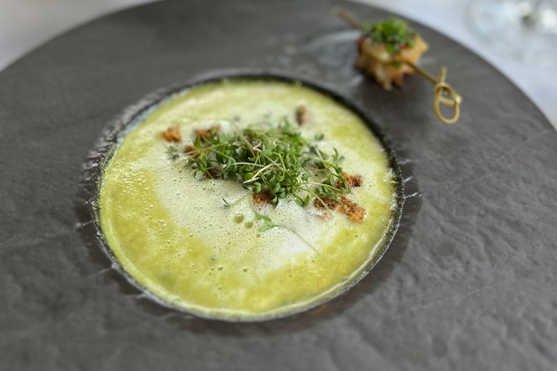 Eine Schale mit grüner Cremesuppe, garniert mit frischen Sprossen und Croutons, serviert auf einem dunklen Teller mit einem Spieß mit gegrillten Lebensmitteln und Microgreens an der Seite.