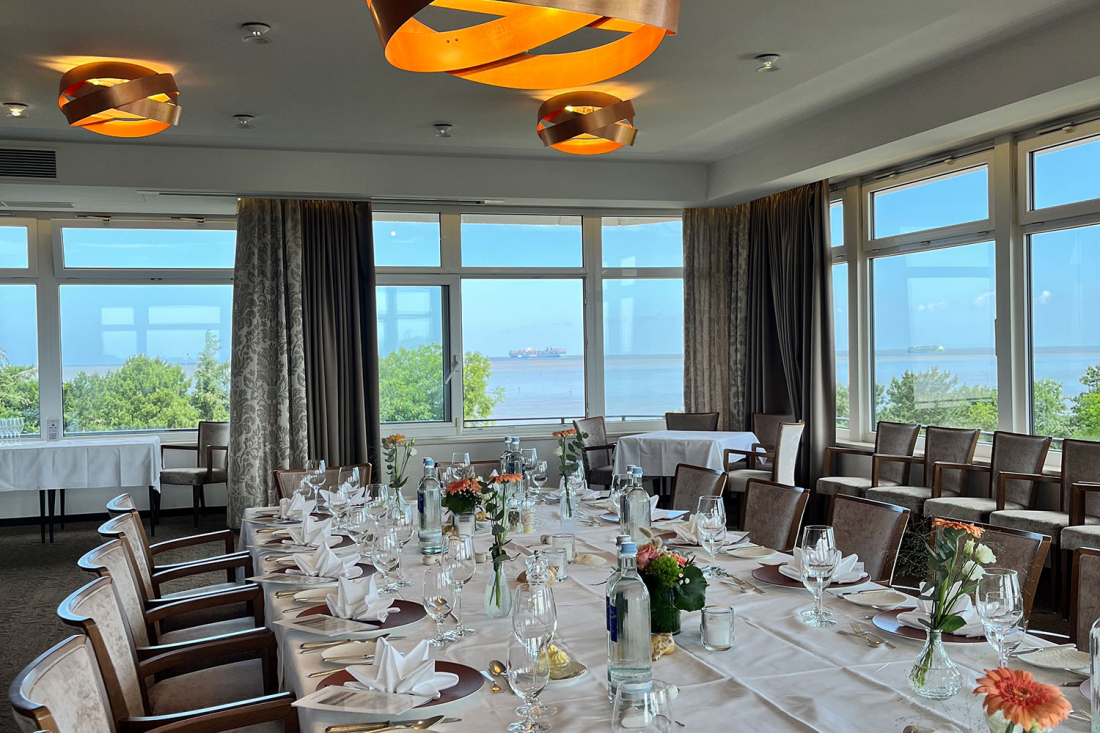 Tagungsraum 2 "Meerblick" mit großen Fenstern mit Blick aufs Meer, einem langen Esstisch mit Tellern, Gläsern, Besteck und Blumenschmuck sowie modernen Hängelampen an der Decke.