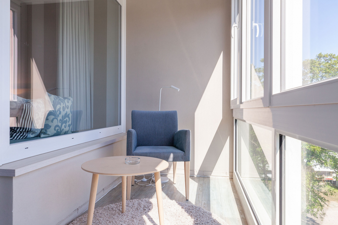 Ein heller, moderner Balkon mit großen Fenstern, einem grauen Sessel, einem kleinen runden Holztisch mit einem Aschenbecher, einer Stehlampe und einem weichen beigen Teppich. Das Sonnenlicht strömt herein und schafft eine gemütliche Atmosphäre.