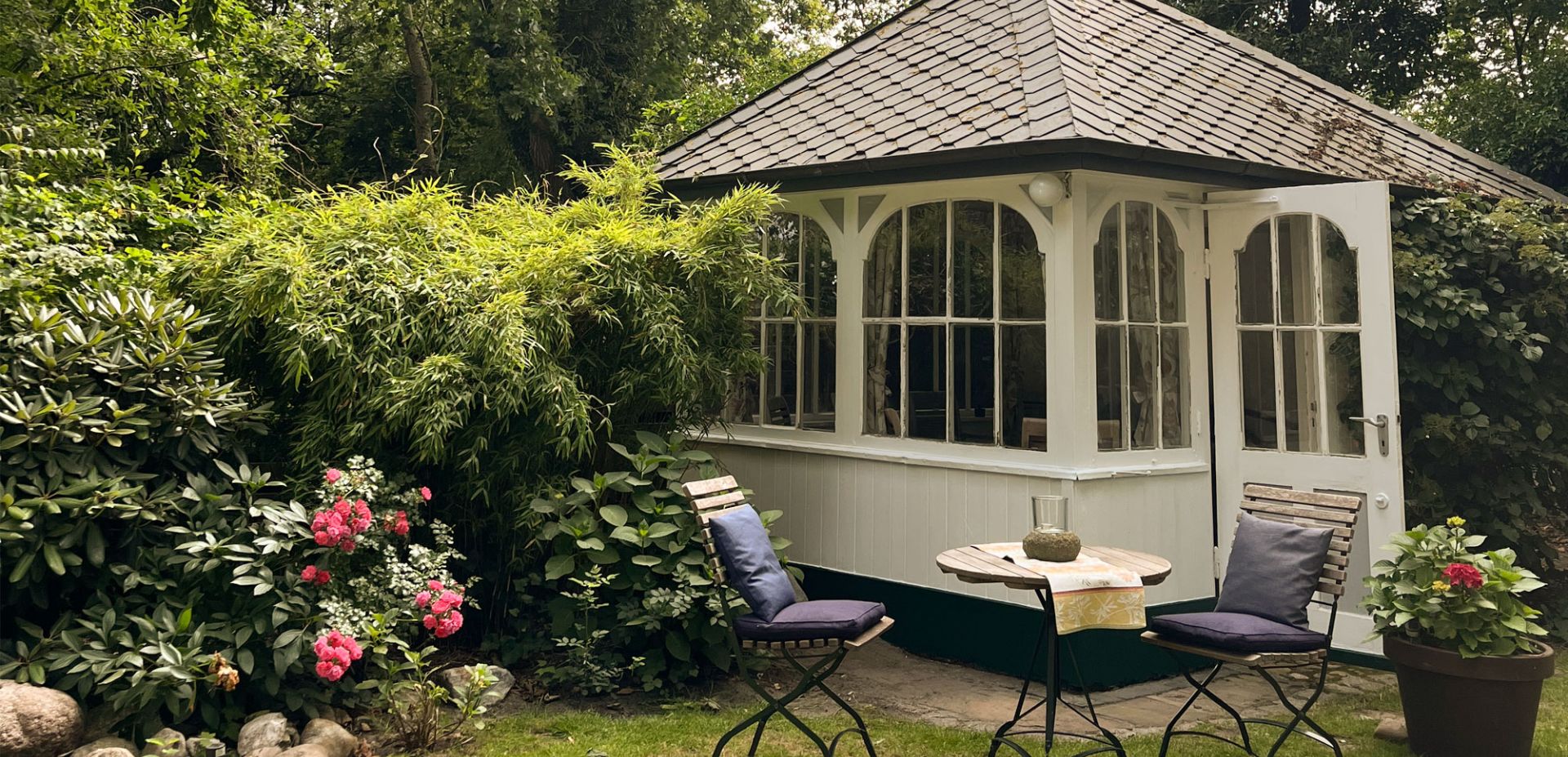 Ein malerischer Gartenpavillon mit großen Fenstern steht umgeben von üppigem Grün, blühenden rosafarbenen Blumen und zwei Holzstühlen mit einem kleinen Tisch auf einem grasbewachsenen Rasen.